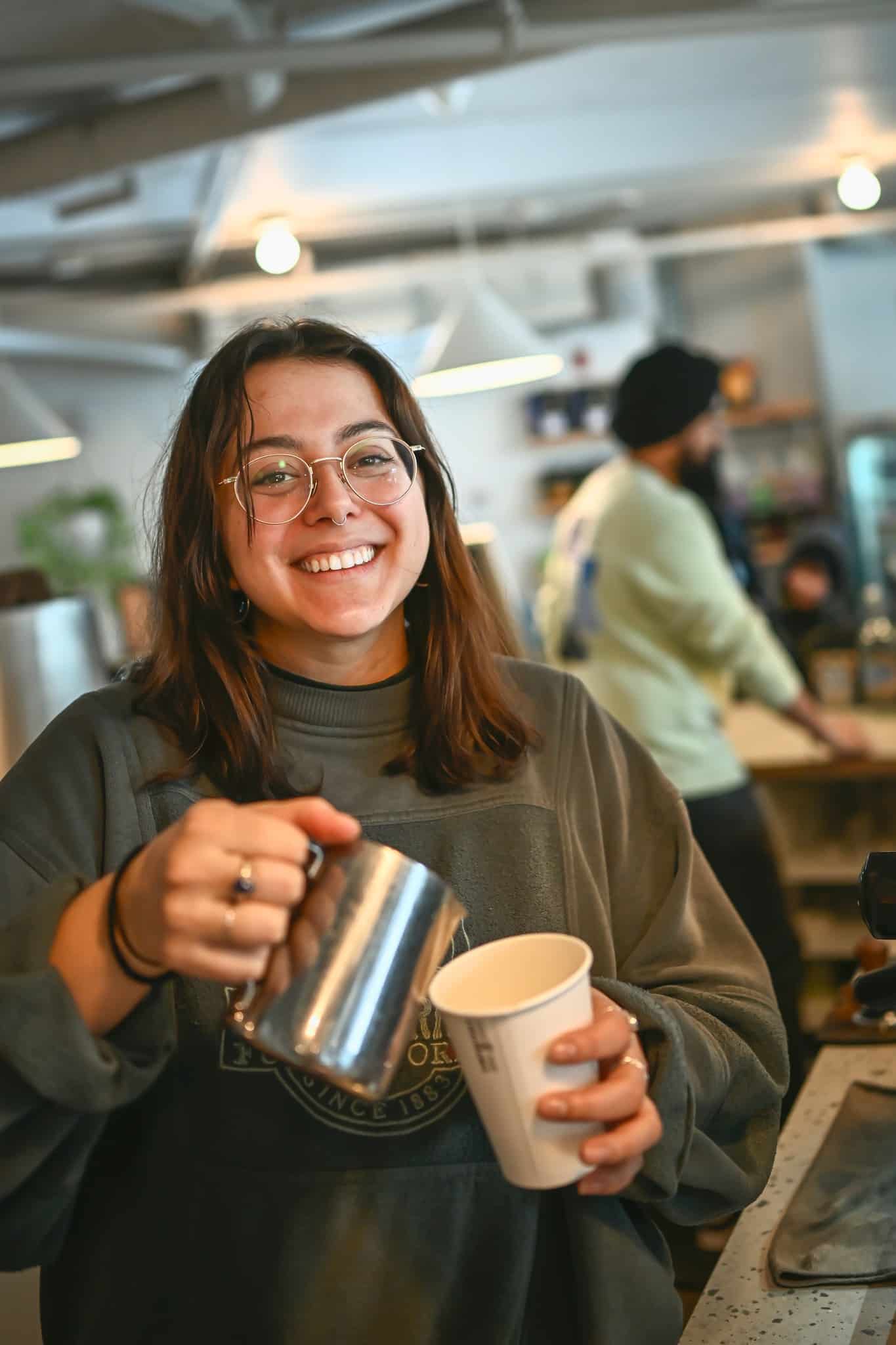 Jeune barista souriante avec des lunettes versant du lait dans une tasse de café dans un café animé.